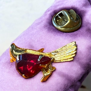 Könnte beinhalten: Goldfarbene Vogelbrosche mit einem großen, herzförmigen, roten Edelstein. Die Flügel des Vogels sind mit strukturierten Linien versehen. Ein separater goldfarbener Nadelverschluss ist ebenfalls sichtbar. Die Brosche befindet sich auf einem lila Hintergrund.