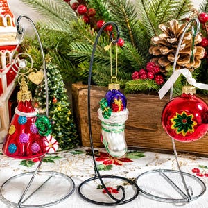 May include: Three glass Christmas ornaments hanging on metal stands. The left is a red cone with colorful glitter dots, the center is a white stocking filled with gifts, and the right is a red sphere with a green and yellow star-shaped design.