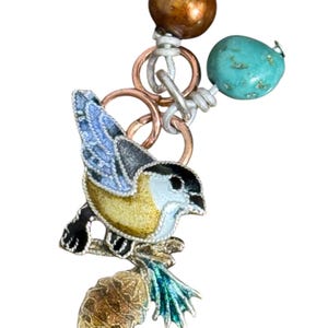 May include: A detailed bird charm with a yellow, black, and blue enamel design, perched on a branch. The charm is connected to a chain with copper rings, a turquoise bead, and a bronze-colored bead. This jewelry piece is suitable for necklaces or bracelets.