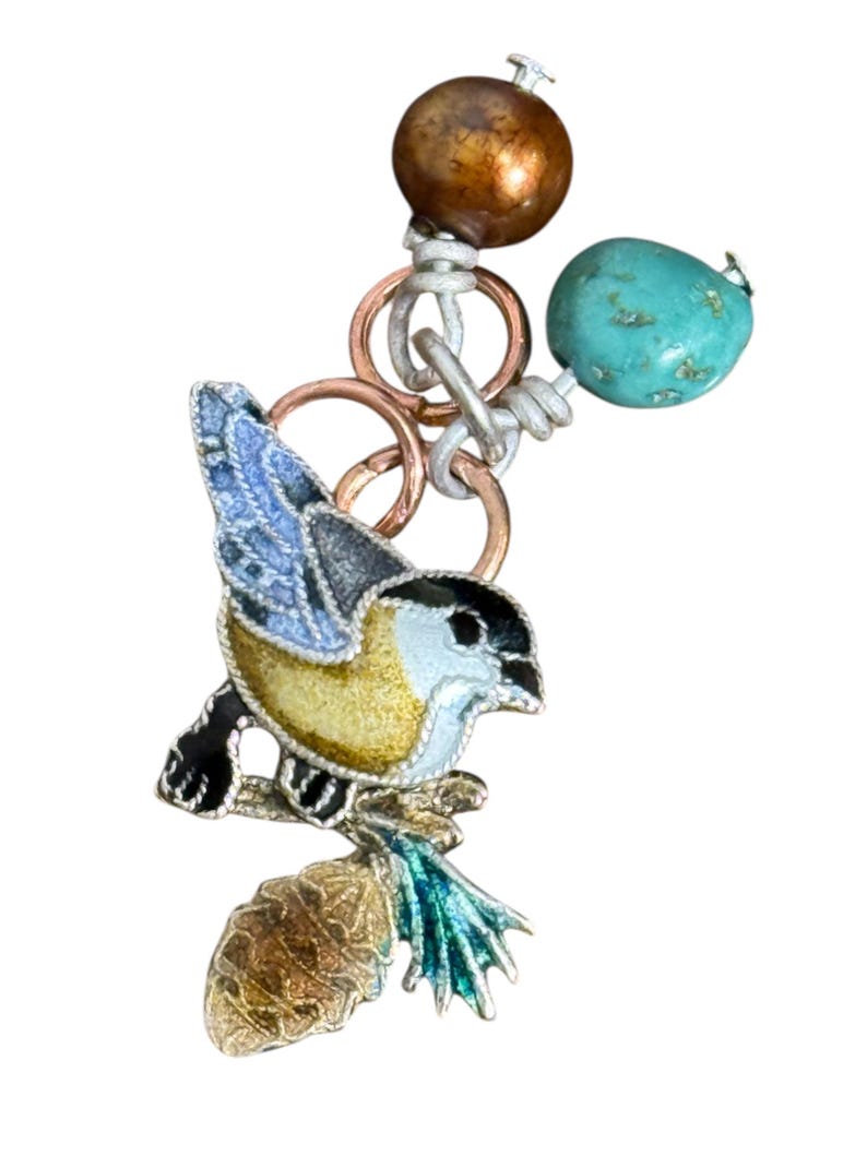 May include: A detailed bird charm with a yellow, black, and blue enamel design, perched on a branch. The charm is connected to a chain with copper rings, a turquoise bead, and a bronze-colored bead. This jewelry piece is suitable for necklaces or bracelets.