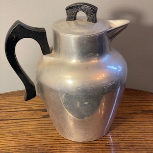 May include: A silver aluminum coffee pot with a black plastic handle and lid. The pot has a rounded body and a spout on the side.