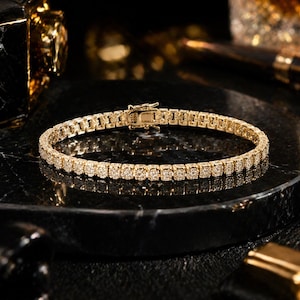 May include: A gold tennis bracelet with numerous small, round, clear gemstones. The bracelet is set on a dark, textured surface, with a gold clasp. The jewelry is displayed in a studio setting with other gold accessories.