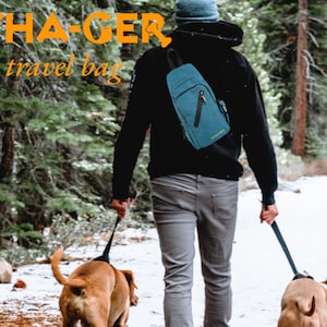 May include: A person walking with two dogs in a snowy forest. The person is wearing a blue hat and a black jacket. They are carrying a blue and black travel bag. The text "WHA-GER Dog travel bag" is visible in the image.