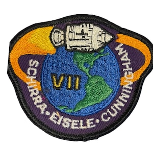 Könnte beinhalten: Gestickter Aufnäher mit einer Rakete über einem blauen Globus mit den Amerikas in Grün. Der Aufnäher hat einen lila Rand mit den Namen SCHIRRA, EISELE und CUNNINGHAM. Die römische Zahl VII ist in Gold.