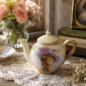 May include: A porcelain teapot with an iridescent cream finish, featuring a painted portrait of a person wearing a purple hat. The teapot has a gold-trimmed lid and a gold handle. It is positioned on a white lace runner next to a floral teacup.