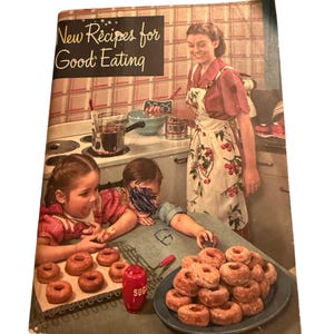 Puede incluir: Portada de libro de cocina vintage titulada "New Recipes for Good Eating". La imagen muestra a una mujer y dos niños en una cocina, con donuts en un plato y una bandeja. También se ve un azucarero rojo.