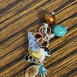 May include: A detailed bird pendant with a yellow, black, and blue enamel design, perched on a brown and green branch. The pendant is connected to a chain with copper rings, a turquoise bead, and a bronze-colored bead. The background is a wooden surface.