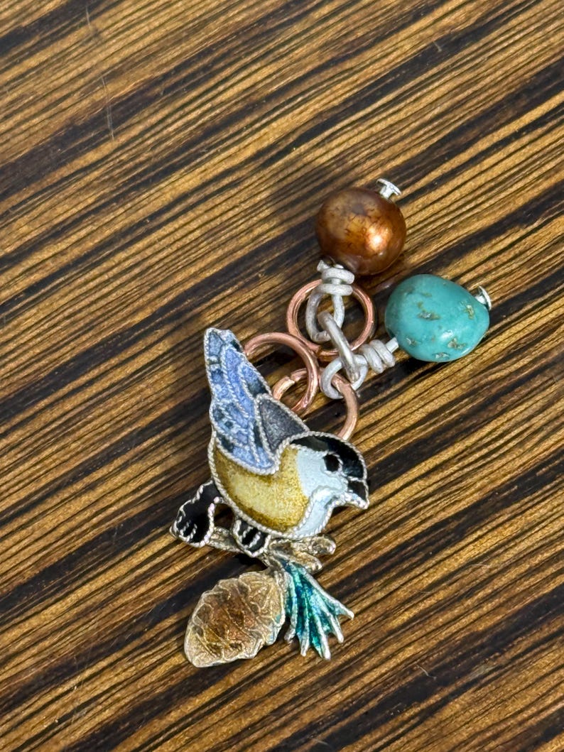 May include: A detailed bird pendant with a yellow, black, and blue enamel design, perched on a brown and green branch. The pendant is connected to a chain with copper rings, a turquoise bead, and a bronze-colored bead. The background is a wooden surface.