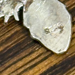 May include: Close-up of a silver-colored metal pendant with a textured surface. The pendant has an irregular shape and appears to be a leaf or similar organic form. The background is a wooden surface with a striped pattern.