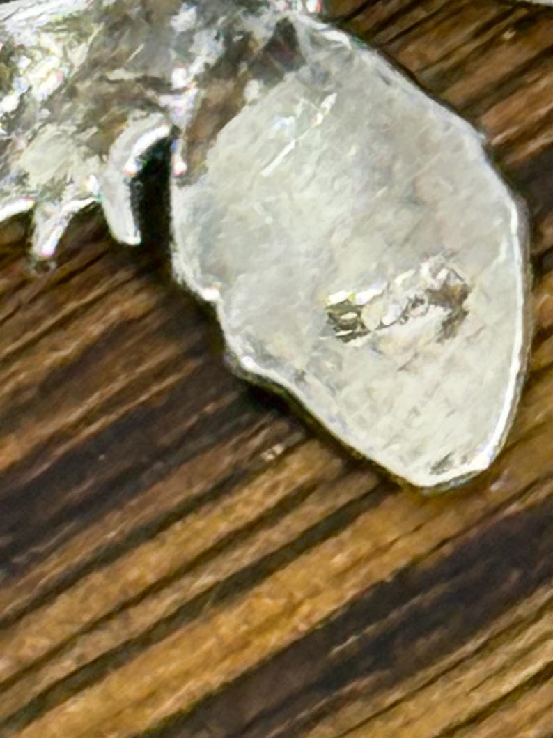 May include: Close-up of a silver-colored metal pendant with a textured surface. The pendant has an irregular shape and appears to be a leaf or similar organic form. The background is a wooden surface with a striped pattern.