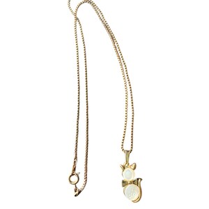 May include: Gold-toned necklace featuring a cat pendant with a pearl-like body and a bow. The chain is a delicate box chain style. The necklace is displayed against a white background.