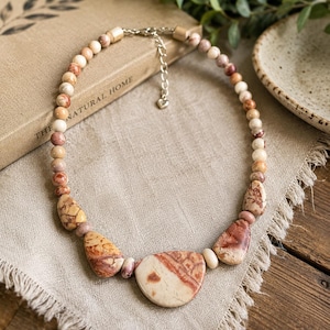 May include: A beaded necklace featuring round, tan, and reddish-brown stone beads that transition into larger, irregular, polished stone pendants at the center. The necklace includes a silver-toned metal chain extender with a small heart-shaped charm.