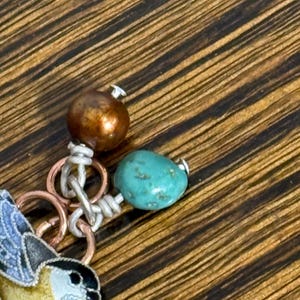 May include: Close-up of a handcrafted jewelry piece featuring a small bird charm with blue and yellow enamel detailing. The piece includes a turquoise bead, a copper-colored bead, and silver-toned metal rings and wire.