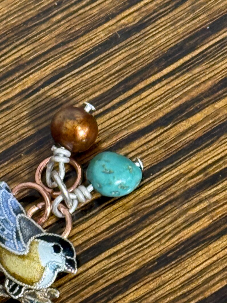 May include: Close-up of a handcrafted jewelry piece featuring a small bird charm with blue and yellow enamel detailing. The piece includes a turquoise bead, a copper-colored bead, and silver-toned metal rings and wire.