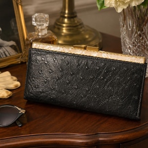 May include: A rectangular black clutch wallet with a textured ostrich-skin pattern. The wallet features a gold-toned metal frame along the top edge, accented with a white pearlescent inlay and a rectangular clasp closure mechanism.
