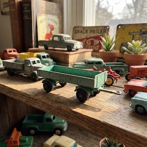 May include: A collection of vintage toy trucks and cars in various colors, including green, red, and yellow. The toys are arranged on a wooden shelf, with a green trailer truck in the foreground. Other items include books, signs, and small potted plants.