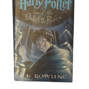 May include: A hardcover book titled "Harry Potter and the Order of the Phoenix" by J.K. Rowling. The cover features an illustration of Harry Potter with glasses, and the title is in a light blue font.