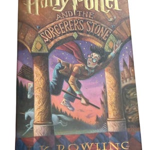 May include: A hardcover book titled Harry Potter and the Sorcerer's Stone by J.K. Rowling. The cover features a colorful illustration of a young person with glasses flying on a broomstick through a stone archway, with a unicorn in the background.
