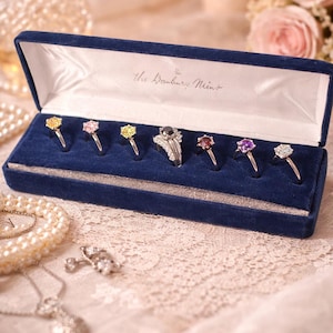 May include: A navy blue velvet jewelry box displaying several gemstone rings. The rings feature various colored stones, including yellow, pink, and purple. The box is lined with white satin and has the text "The Danbury Mint" printed on the inside.