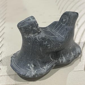 May include: A small, gray stone sculpture of a bird with intricate carvings. The bird is perched on a base with a carved design.