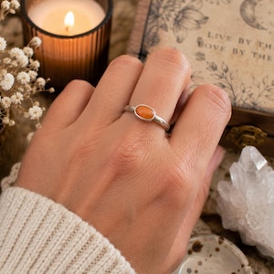 May include: A silver ring featuring a smooth, oval-shaped orange stone set in a simple metal band. The ring is worn on a finger, showcasing the polished finish of the silver and the vibrant, solid color of the gemstone centered on the band.