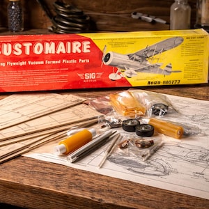 May include: A vintage model airplane kit with the name "CUSTOMAIRE" on the box. The box is red and yellow, and the kit includes various parts, blueprints, and tools. The kit is set on a wooden surface.
