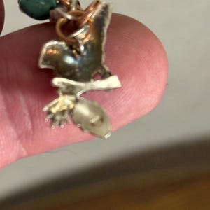 May include: A handcrafted silver-toned pendant with a bird-like design, featuring copper wire accents and a small, green bead. The pendant has a unique, organic shape and a polished finish, showcasing intricate details.