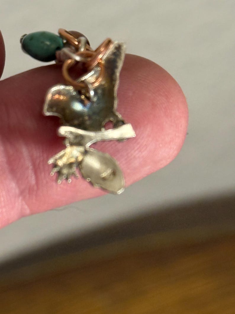 May include: A handcrafted silver-toned pendant with a bird-like design, featuring copper wire accents and a small, green bead. The pendant has a unique, organic shape and a polished finish, showcasing intricate details.