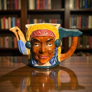Può includere: Una teiera in ceramica con un design da capo nativo americano. La teiera presenta un volto dettagliato con un copricapo, piume e un manico marrone. I colori includono marrone, giallo, blu e rosso.