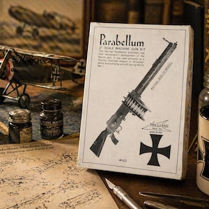Könnte beinhalten: Eine Vintage 2" Parabellum Maschinengewehr-Bausatzbox mit einer Schwarz-Weiß-Illustration der Waffe. Die Box befindet sich neben einem Modellflugzeug, Farbtöpfen und einer Tasse mit einem schwarzen Kreuzdesign. Der Text auf der Box lautet "Parabellum".