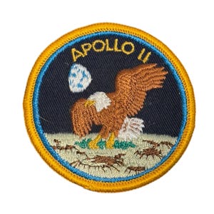 May include: Embroidered patch featuring an eagle with outstretched wings, a moon, and the text "APOLLO II." The patch has a blue background with a yellow border and depicts a scene with crabs and grass.