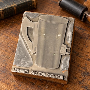 May include: A rectangular metal printing block mounted on a wooden base, featuring an embossed graphic of a mug with an electrical plug and the text HERSHEY'S HOT CUP STDR-N2 engraved along the bottom edge of the metal surface.