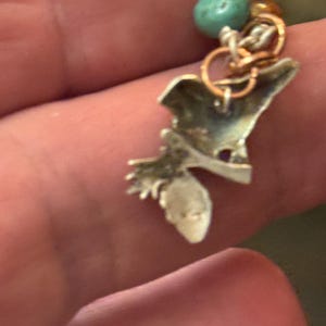 May include: A silver-toned charm with a turquoise bead and small gold and silver rings. The charm has an irregular shape with a small, pointed extension. The charm is attached to a small silver ring.