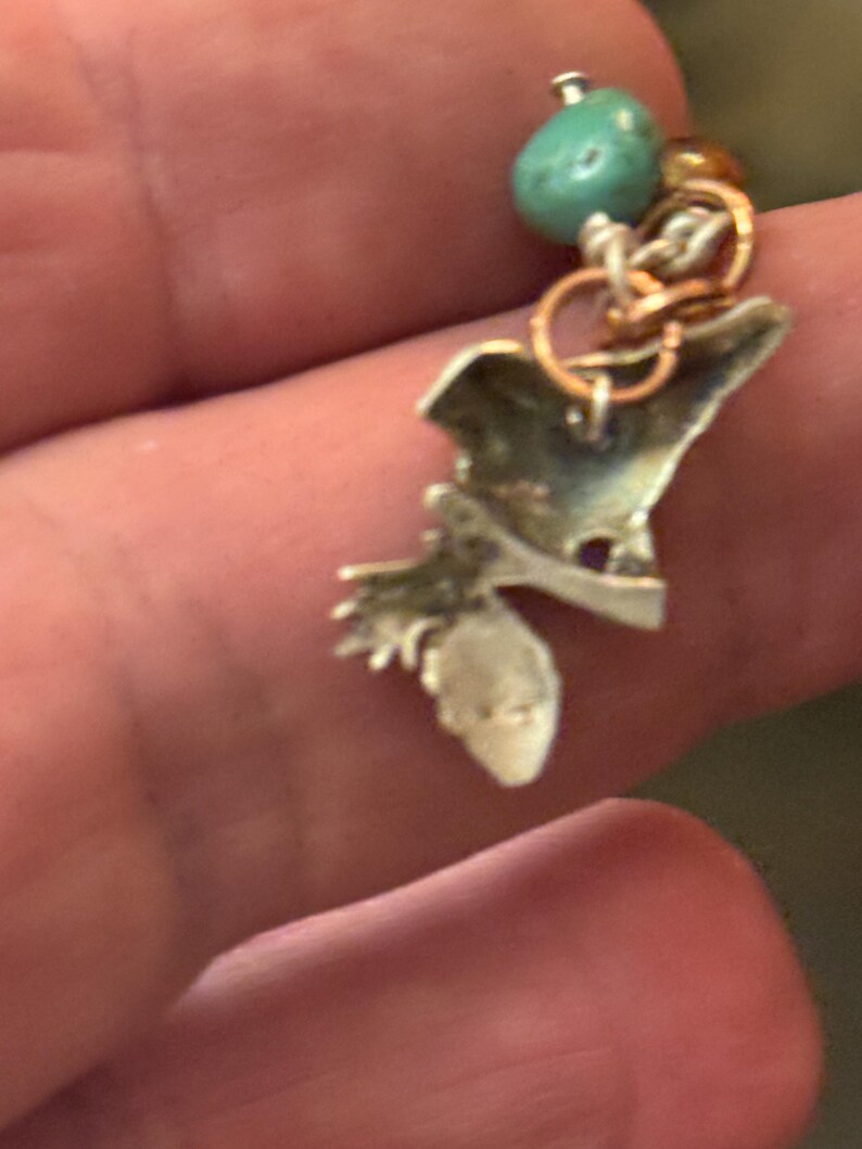 May include: A silver-toned charm with a turquoise bead and small gold and silver rings. The charm has an irregular shape with a small, pointed extension. The charm is attached to a small silver ring.