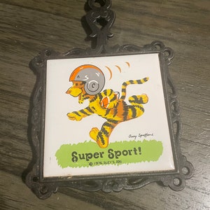 May include: A black cast iron trivet with a white ceramic tile featuring a cartoon tiger wearing a helmet and running. The tile has the text "Super Sport!" and "© 1976 Suzy's Zoo" printed on it.