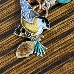 May include: A detailed pendant featuring a colorful bird perched on a branch with a pinecone. The bird has a yellow body, blue wings, and a black head. The pendant is attached to a chain with turquoise beads and copper and silver-toned rings.