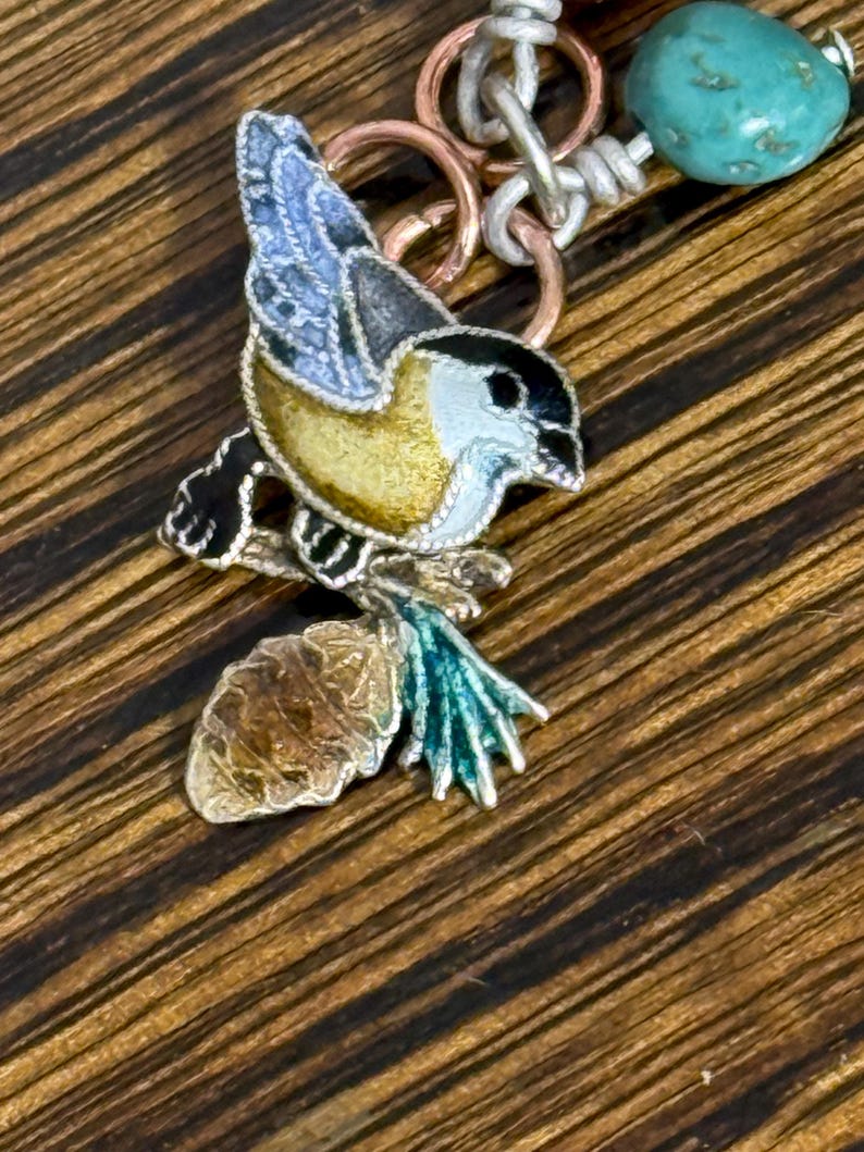 May include: A detailed pendant featuring a colorful bird perched on a branch with a pinecone. The bird has a yellow body, blue wings, and a black head. The pendant is attached to a chain with turquoise beads and copper and silver-toned rings.