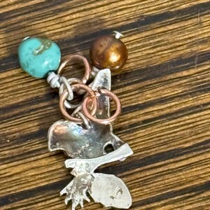 May include: A handcrafted pendant featuring a silver-toned metal design with a leaf and bird-like shape. It is adorned with a turquoise bead and a bronze-colored bead, connected by silver and copper-colored rings. The pendant is designed for jewelry.