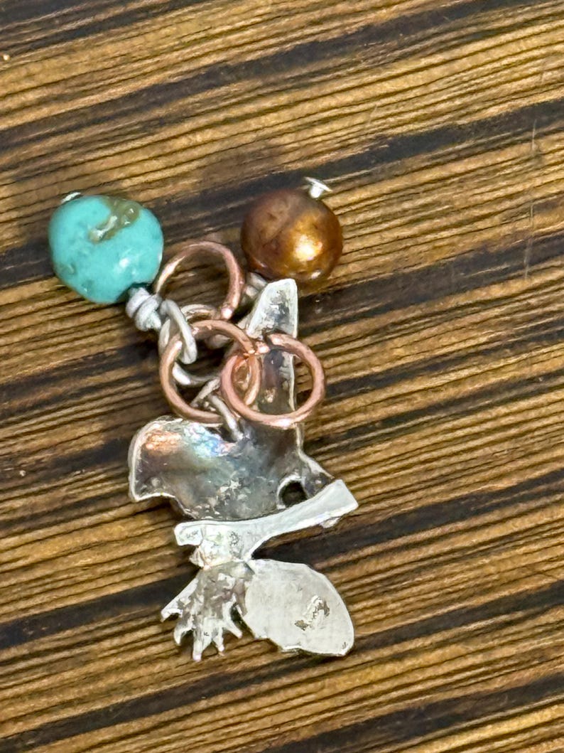 May include: A handcrafted pendant featuring a silver-toned metal design with a leaf and bird-like shape. It is adorned with a turquoise bead and a bronze-colored bead, connected by silver and copper-colored rings. The pendant is designed for jewelry.