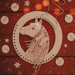 K&ouml;nnte beinhalten: Weihnachts-Adventskalender mit h&ouml;lzernem Pferdekopf, rosa Garnm&auml;hne und Schleife. Der Pferdekopf befindet sich in einem Holzring mit kleinen L&ouml;chern. Enth&auml;lt nummerierte Holzkreise und Schneeflocken auf rotem Hintergrund.