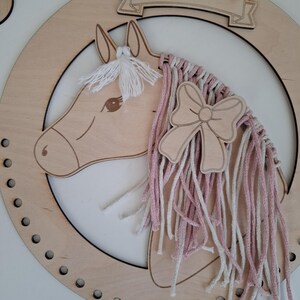 K&ouml;nnte beinhalten: Wanddekoration mit h&ouml;lzernem Pferdekopf in hellbrauner Ausf&uuml;hrung. Das Pferd hat eine M&auml;hne aus wei&szlig;em Garn und einen Zopf aus rosa und wei&szlig;em Garn. Eine Holzschleife ist an dem Zopf befestigt. Das Pferd befindet sich in einem Holzkreis mit einem Banner oben.