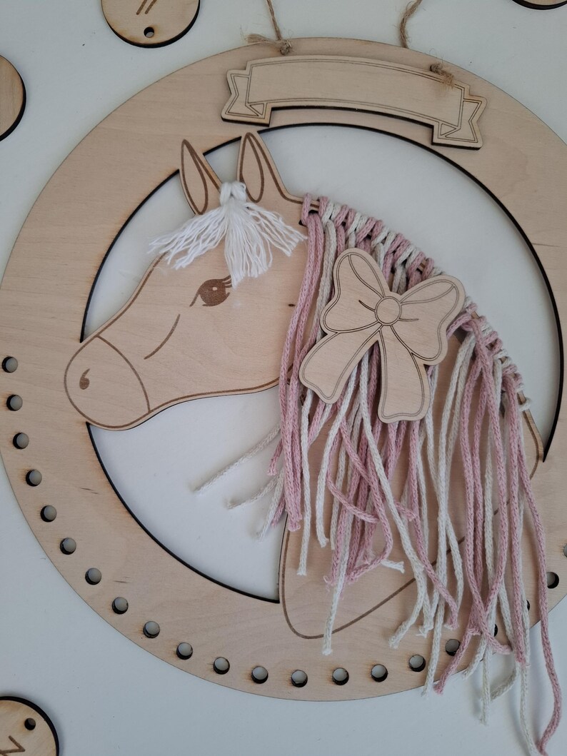 K&ouml;nnte beinhalten: Wanddekoration mit h&ouml;lzernem Pferdekopf in hellbrauner Ausf&uuml;hrung. Das Pferd hat eine M&auml;hne aus wei&szlig;em Garn und einen Zopf aus rosa und wei&szlig;em Garn. Eine Holzschleife ist an dem Zopf befestigt. Das Pferd befindet sich in einem Holzkreis mit einem Banner oben.