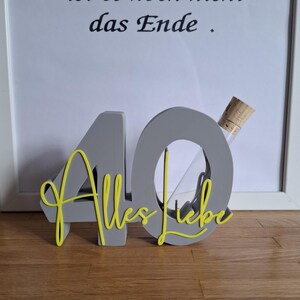Könnte beinhalten: Eine graue Holz-40 mit gelbem Schriftzug 'Alles Liebe'. Eine kleine Glasampulle mit Korken ist in die '0' eingesetzt. Der Hintergrund enthält einen gerahmten Druck mit Text.