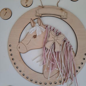 K&ouml;nnte beinhalten: Wanddekoration mit h&ouml;lzernem Pferdekopf, rosa-wei&szlig;em Garnm&auml;hne und Schleife. Das Pferd befindet sich in einem Holzkreis mit einem Banner oben. Enth&auml;lt Holznummernschilder 1, 3, 4 und ein paar andere.