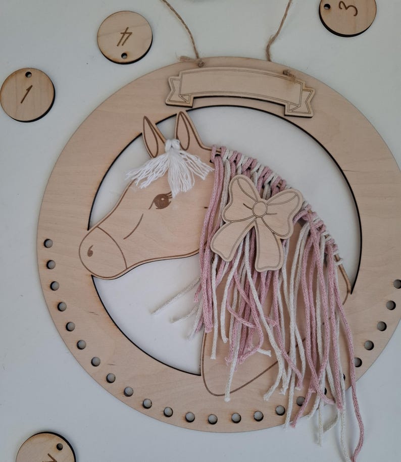 K&ouml;nnte beinhalten: Wanddekoration mit h&ouml;lzernem Pferdekopf, rosa-wei&szlig;em Garnm&auml;hne und Schleife. Das Pferd befindet sich in einem Holzkreis mit einem Banner oben. Enth&auml;lt Holznummernschilder 1, 3, 4 und ein paar andere.