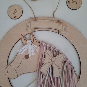 K&ouml;nnte beinhalten: Wanddekoration aus Holz in Form eines Pferdekopfes mit einer M&auml;hne aus rosa und wei&szlig;em Garn und einer Holzschleife. Das Pferd befindet sich in einer hufeisenf&ouml;rmigen Holzform. Enth&auml;lt zwei Holzkreise mit den Zahlen 3 und 4.