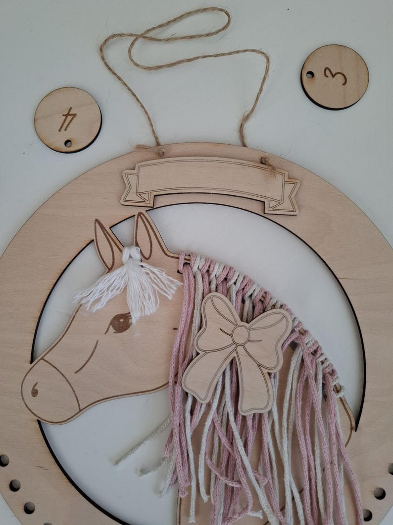 K&ouml;nnte beinhalten: Wanddekoration aus Holz in Form eines Pferdekopfes mit einer M&auml;hne aus rosa und wei&szlig;em Garn und einer Holzschleife. Das Pferd befindet sich in einer hufeisenf&ouml;rmigen Holzform. Enth&auml;lt zwei Holzkreise mit den Zahlen 3 und 4.
