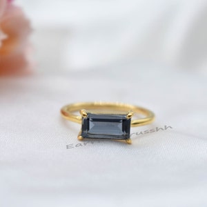 May include: A gold ring with a dark gray rectangular gemstone. The ring is on a white fabric background.