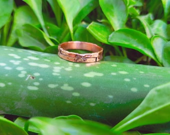 Op de afbeelding: Een gehamerde koperen ring met een getextureerd oppervlak. De ring staat tegen een achtergrond van groene bladeren en een groen, gespikkeld oppervlak. Het ontwerp van de ring heeft een gespleten band met een gehamerde textuur, wat een rustieke esthetiek laat zien.