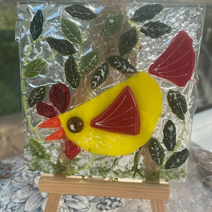 May include: A yellow glass bird with red accents is perched on a leafy branch. The bird is made of stained glass and has a textured, cracked glass background.